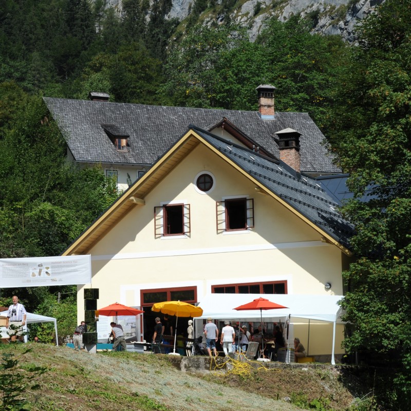 Mehrere Personen unter bunten Sonnenschirmen vor einem hellen Gebäude mit steilem Dach, im Hintergrund reich bewaldete
                                 Hügel. (KI-generierter Alt-Text, erstellt mit GPT-4.1-mini)