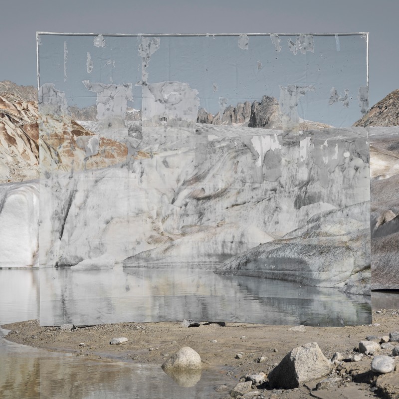 Large rectangular mirror stands by a glacier lake, reflecting rugged rocks and ice in the background. (AI-generated
                                 alt-text, generated with GPT-4.1-mini)