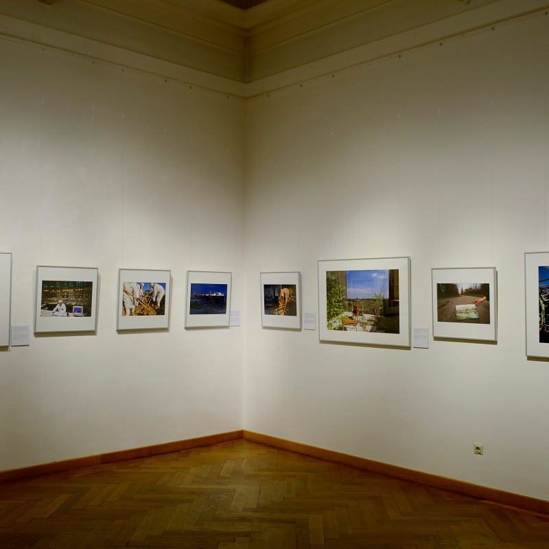 Gallery corner with nine framed color photographs on white walls and parquet floor. (AI-generated alt-text, generated
                                 with GPT-4.1-mini)