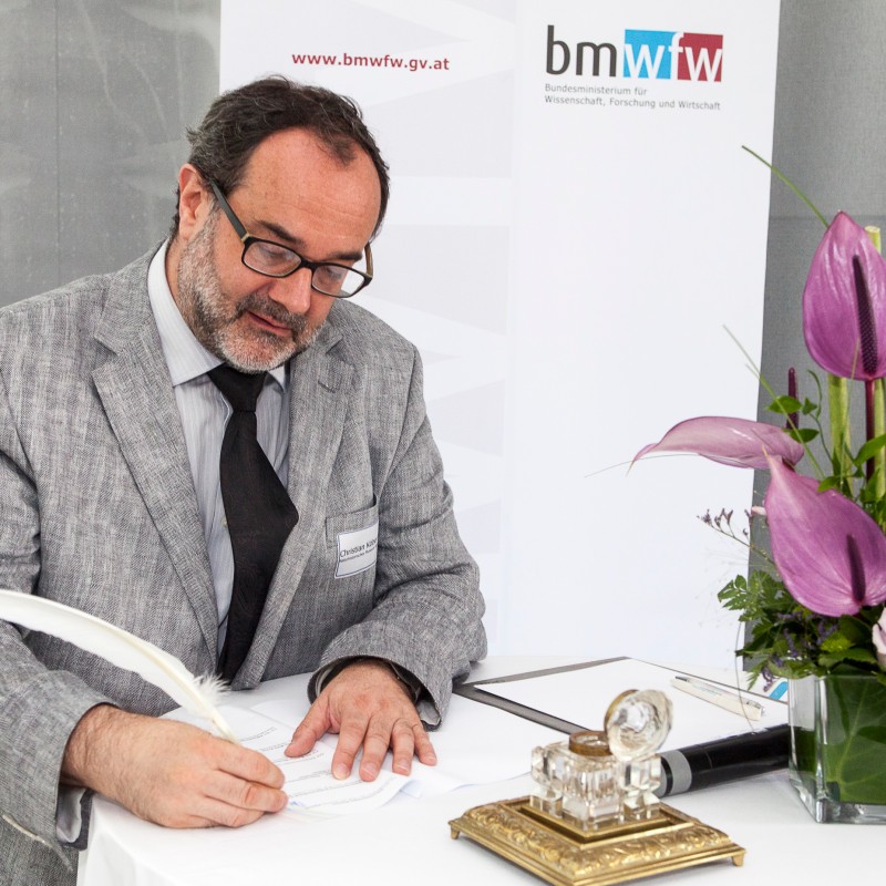 Man with glasses, gray jacket, and striped tie sits at a table, writing with a quill on paper. Purple flower arrangement
                                 beside him. (AI-generated alt-text, generated with GPT-4.1-mini)