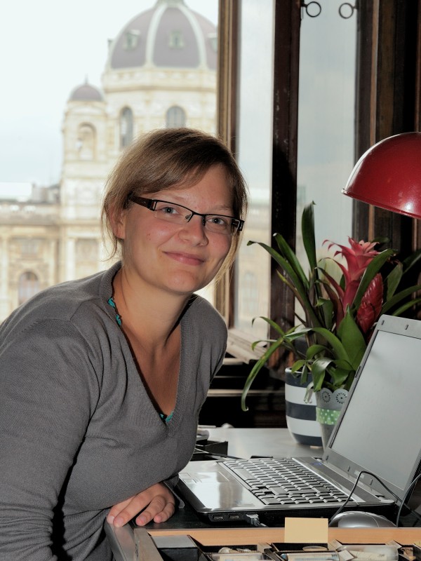 Frau mit Brille und grauem Pullover sitzt an einem Schreibtisch mit geöffnetem Laptop vor einem Fenster, im Hintergrund
                                 ein historisches Kuppelgebäude. (KI-generierter Alt-Text, erstellt mit GPT-4.1-mini)