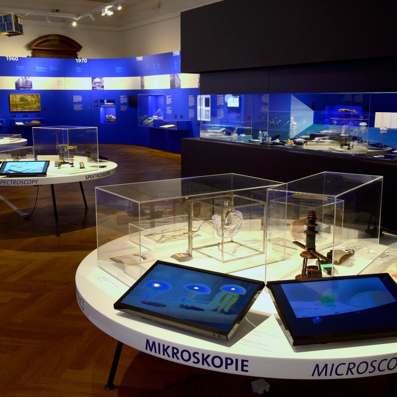 Exhibition
                                 with several round tables displaying historical scientific instruments and books in glass cases. Blue wall panels with exhibits
                                 and descriptions in the background. (AI-generated alt-text, generated with GPT-4.1-mini)