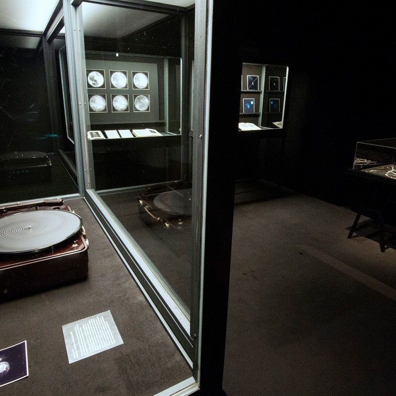 Dimly lit exhibition room with illuminated display cases, including a record player in a suitcase and several framed
                                 items on the walls. (AI-generated alt-text, generated with GPT-4.1-mini)