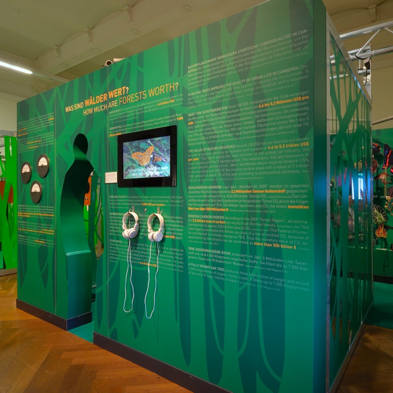 Grüne Ausstellungswände mit aufgedrucktem Text, Monitoren und Kopfhörern in einem Museumsraum. (KI-generierter Alt-Text,
                                 erstellt mit GPT-4.1-mini)