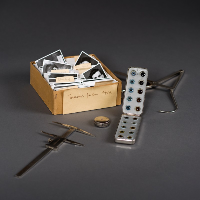 Brauner Karton mit schwarz-weißen Porträtfotos, Metallbox mit mehreren Glasobjekten, Messschieber, Zirkel, kleine
                                 Dose und weiteres Metallinstrument auf grauem Untergrund. (KI-generierter Alt-Text, erstellt mit GPT-4.1-mini)