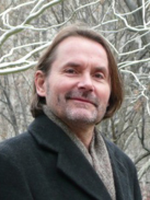 Mann mittleren Alters mit halblangen braunen Haaren und Bart, lächelt leicht, trägt dunklen
                                 Mantel und Schal. Unscharfe, winterliche Äste im Hintergrund. (KI-generierter Alt-Text, erstellt mit GPT-4.1-mini)