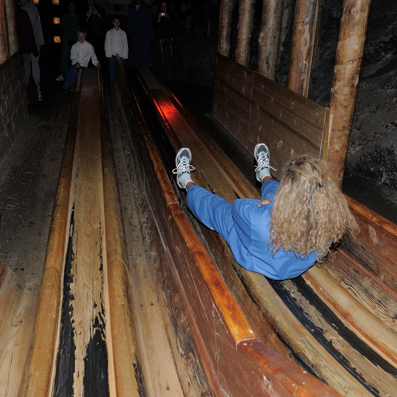 Person with curly blond hair slides down wooden chutes between timber beams. Two other people stand at the end of
                                 the chutes. (AI-generated alt-text, generated with GPT-4.1-mini)