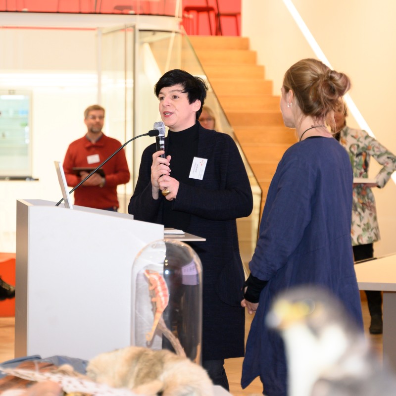 Woman at lectern speaking into a microphone, another woman standing beside her; multiple people seated and watching
                                 in the background. (AI-generated alt-text, generated with GPT-4.1-mini)