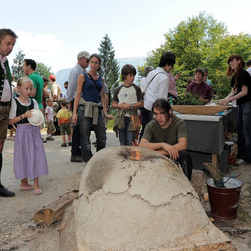 Multiple people in casual clothing
                                 observe a historic clay oven outdoors. A girl in traditional dress stands next to a man in folk costume. In the background,
                                 more people work at wooden tables. (AI-generated alt-text, generated with GPT-4.1-mini)