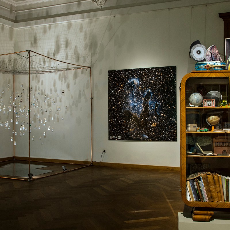Museum exhibit with a display cabinet full of books, globes, rock samples, and models on the right, and an art installation
                                 with a large starry sky photo on the left. (AI-generated alt-text, generated with GPT-4.1-mini)