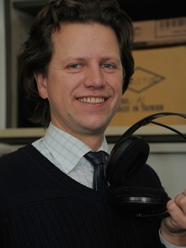 Mann mit mittellangem, gelocktem Haar, Hemd, Pullover und Krawatte hält Kopfhörer in der rechten Hand und lächelt
                                 in die Kamera. (KI-generierter Alt-Text, erstellt mit GPT-4.1-mini)