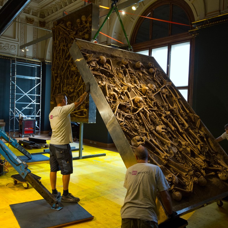 Two men in work shorts and museum shirts position a steeply angled slab with numerous human skeletons under a domed
                                 ceiling. (AI-generated alt-text, generated with GPT-4.1-mini)