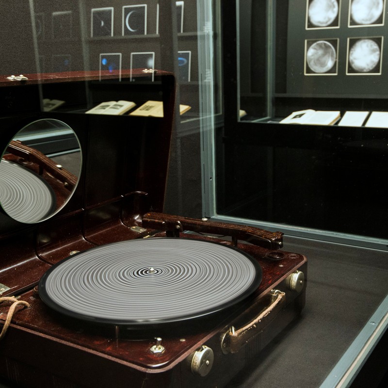 Brown record player with open lid
                                 and viewing window, visible grooved platter, framed black-and-white images on the wall behind. (AI-generated alt-text, generated
                                 with GPT-4.1-mini)