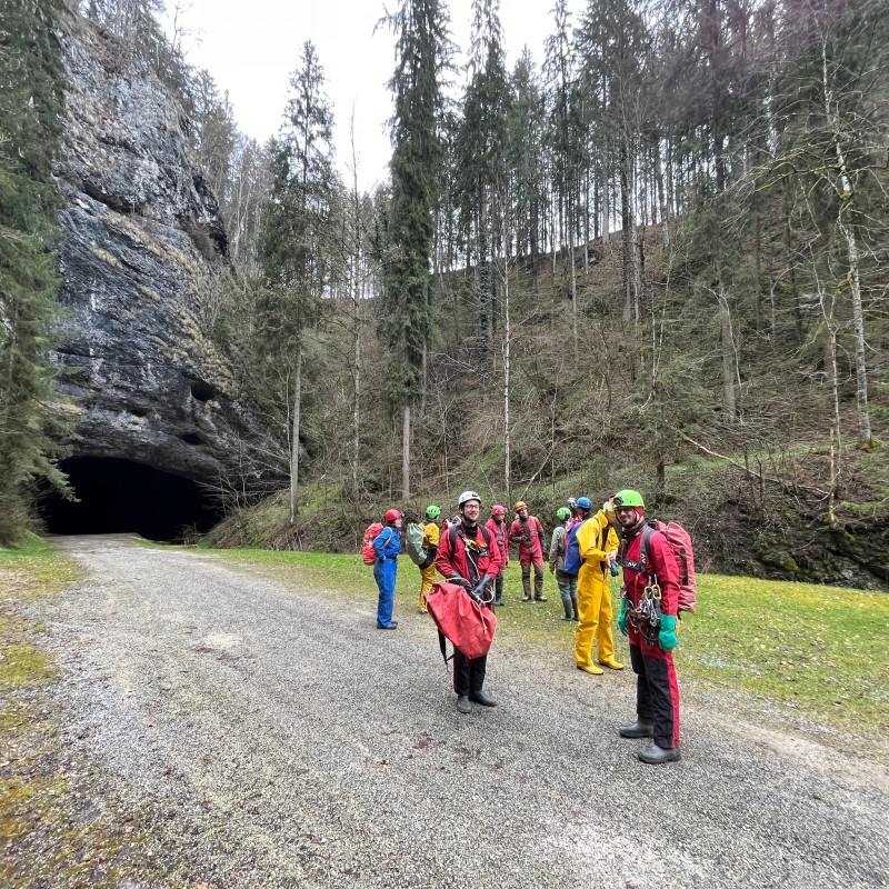 Gruppe von Höhlenforschern in Schutzkleidung steht auf einem Weg vor dem dunklen Höhleneingang der Lurgrotte Semriach,
                                 umgeben von steilem Felsen und Nadelwald. (KI-generierter Alt-Text, erstellt mit GPT-4.1-mini)