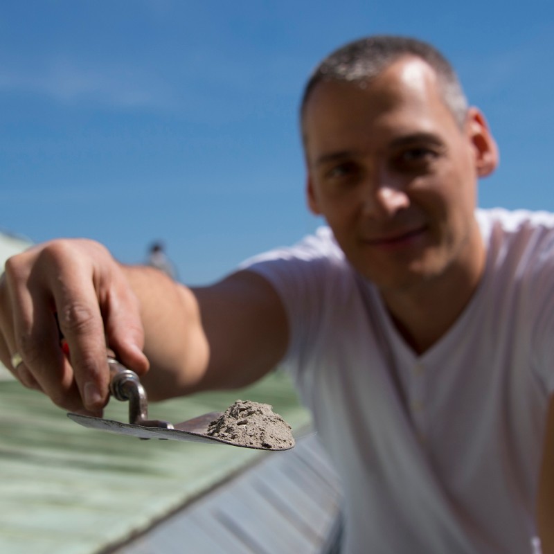 Man in a white T-shirt holds a trowel with gray material toward the camera, rooftop and blue sky in the background.
                                 (AI-generated alt-text, generated with GPT-4.1-mini)