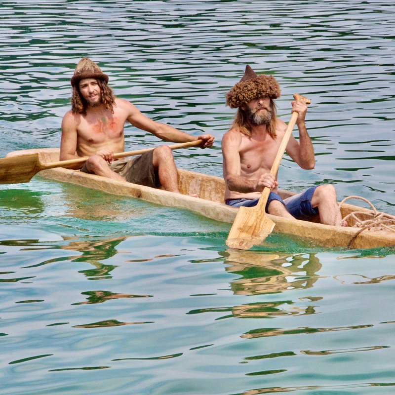 Two men with long hair and beards, wearing traditional headgear, bare-chested and holding oars, in a hand-carved wooden boat on the water