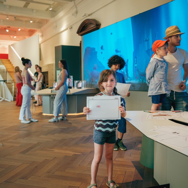 Child with short hair holds a sheet of paper; in the background, exhibition room with several people and large screen
                                 showing underwater scene. (AI-generated alt-text, generated with GPT-4.1-mini)