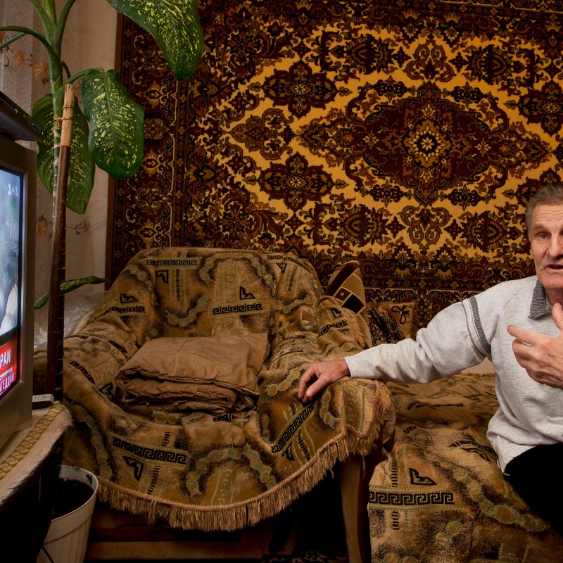 Older man with short hair sits in an armchair in front of a patterned wall tapestry, gesturing with his right hand.
                                 A television showing news and flames is visible on the left. (AI-generated alt-text, generated with GPT-4.1-mini)