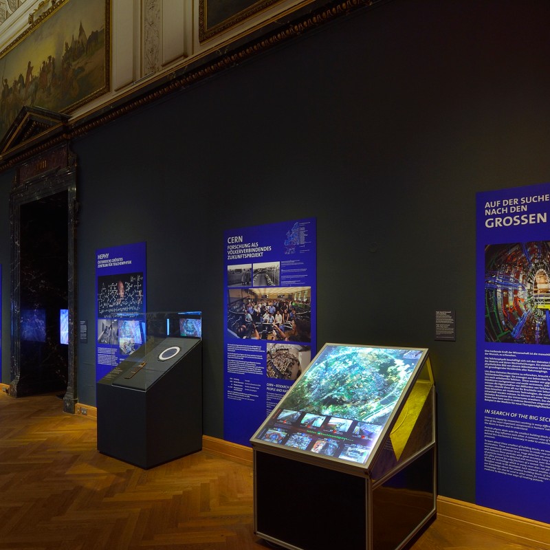 Blick
                                 in einen Ausstellungsraum mit mehreren beleuchteten Vitrinen und großformatigen, blau-gelben Informationstafeln an den Wänden.
                                 (KI-generierter Alt-Text, erstellt mit GPT-4.1-mini)