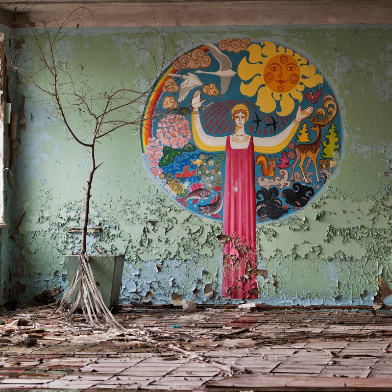 Abandoned room with peeling green walls, a bare tree in a bucket, and a colorful mural featuring a sun, a woman,
                                 animals, and a rainbow. (AI-generated alt-text, generated with GPT-4.1-mini)