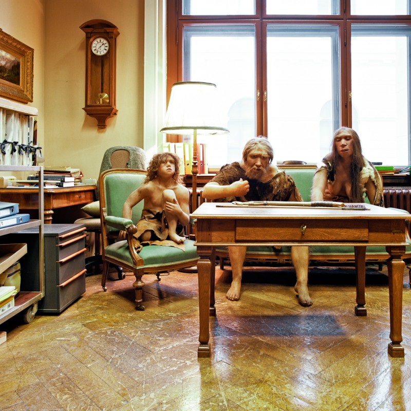 Museumsszene mit drei nachgestellten Neandertalern in einfacher Kleidung, sitzend an einem Holztisch in einem Raum
                                 mit großen Fenstern und Bücherregalen. (KI-generierter Alt-Text, erstellt mit GPT-4.1-mini)