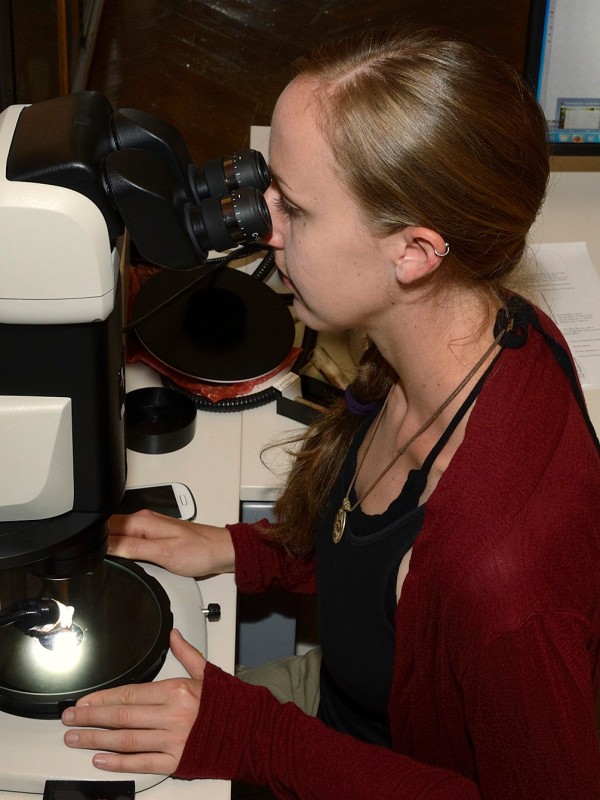 Frau
                                 mit langem Zopf betrachtet durch ein Mikroskop, trägt dunkles Top und roten Cardigan, sitzt an einem Labortisch. (KI-generierter
                                 Alt-Text, erstellt mit GPT-4.1-mini)