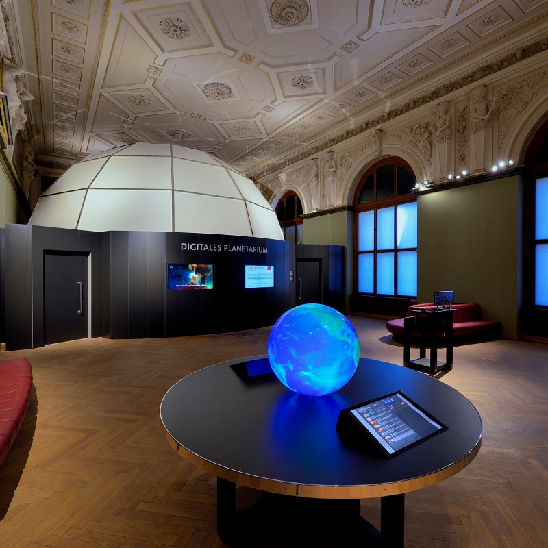 Großer Ausstellungsraum mit Kuppel, runder Projektionsfläche
                                 und leuchtender Weltkugel auf einem Tisch. (KI-generierter Alt-Text, erstellt mit GPT-4.1-mini)