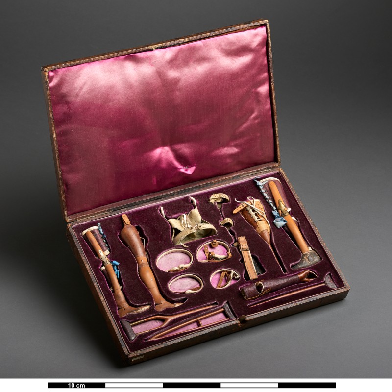 Open case containing medical instruments and prosthetics made of wood and metal, arranged in purple lining. (AI-generated
                                 alt-text, generated with GPT-4.1-mini)