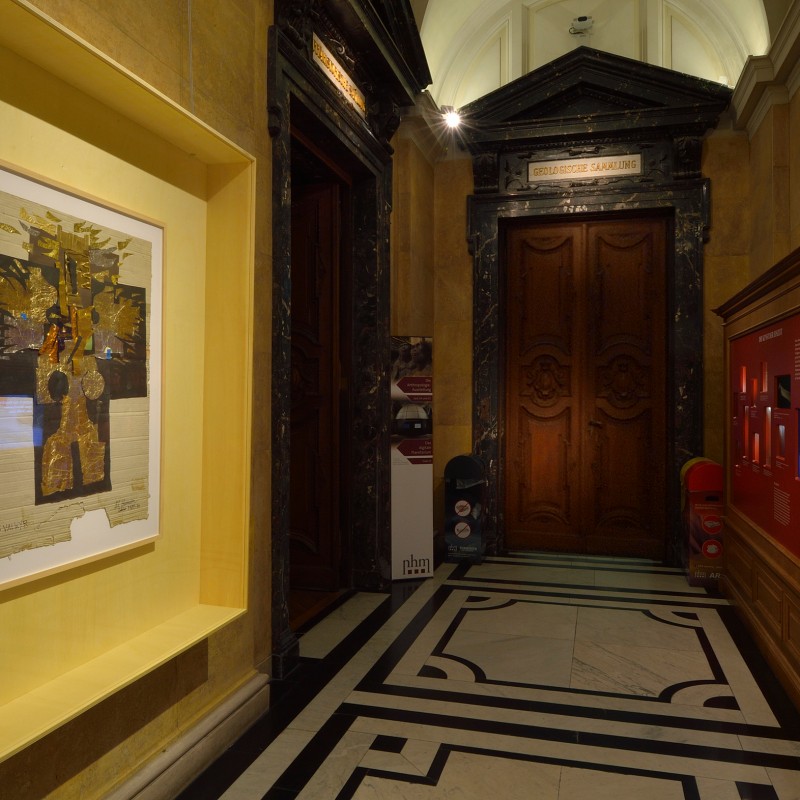 Painting with gold and black shapes in a recessed niche of a museum hallway. On the right, information panels with
                                 red background. (AI-generated alt-text, generated with GPT-4.1-mini)