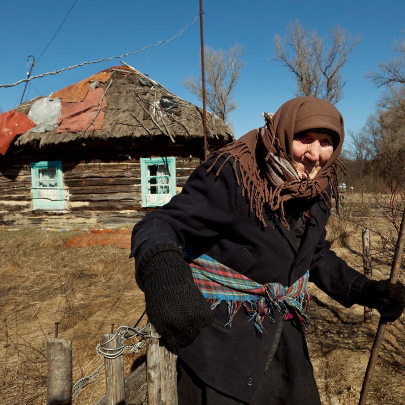 Elderly woman with headscarf and stick stands in the foreground of a rural scene, behind her a small, dilapidated
                                 wooden hut with a damaged roof. (AI-generated alt-text, generated with GPT-4.1-mini)