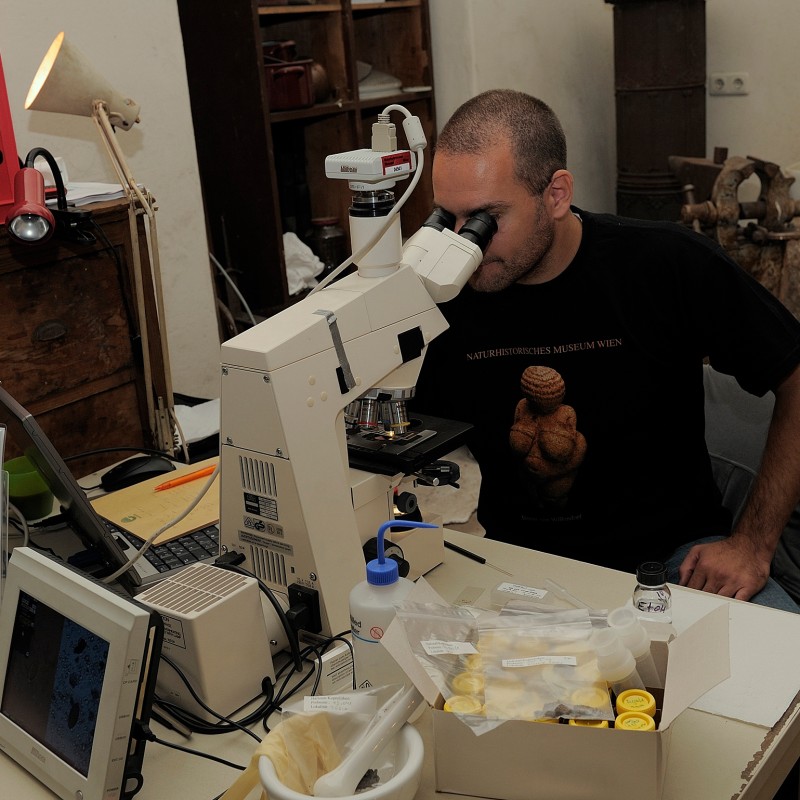 Man with closely cropped hair sits at a microscope and looks through it, surrounded by various lab equipment and
                                 small sample containers. (AI-generated alt-text, generated with GPT-4.1-mini)