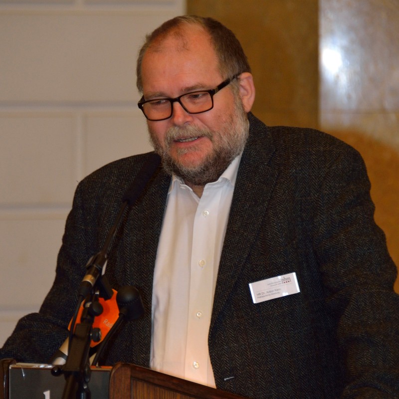 : Mann mit Brille und Bart spricht an einem Rednerpult mit Mikrofon, trägt weißen Hemd und dunklen Blazer. (KI-generierter Alt-Text, erstellt mit GPT-4.1-mini)