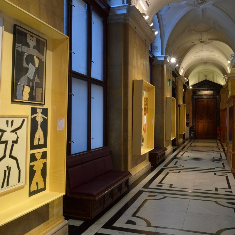 Museum corridor with illuminated display cases containing abstract black-and-white collages or graphics. Ornate
                                 ceiling, tiled floor. (AI-generated alt-text, generated with GPT-4.1-mini)
