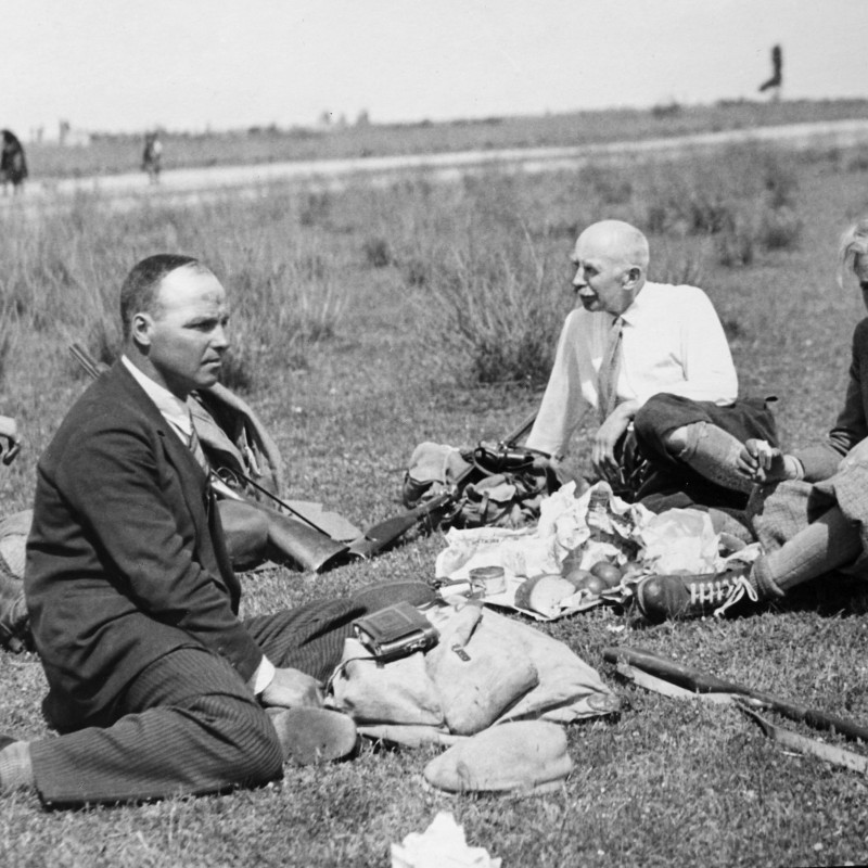 Vier Männer sitzen auf einer Wiese und machen eine Pause mit verstreuten Gegenständen und Essen auf einer Decke.
                                 (KI-generierter Alt-Text, erstellt mit GPT-4.1-mini)