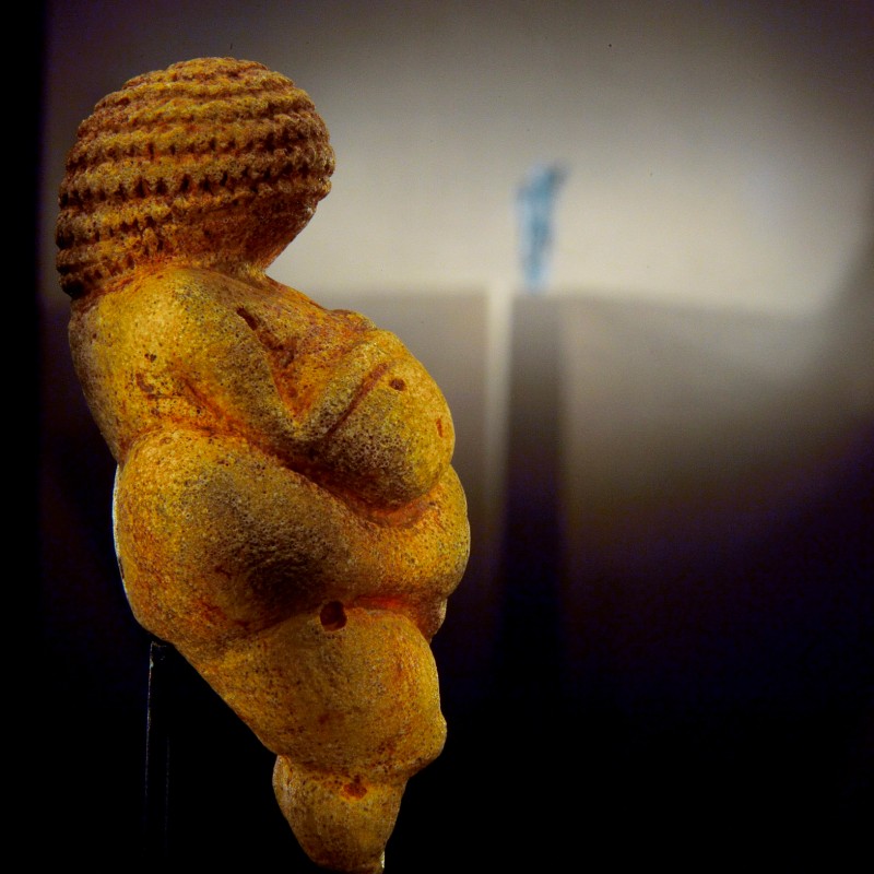 : Close-up of an ochre-colored, corpulent female figurine with detailed hair pattern against a dark background, shadow to the right. (AI-generated alt-text, generated with GPT-4.1-mini)