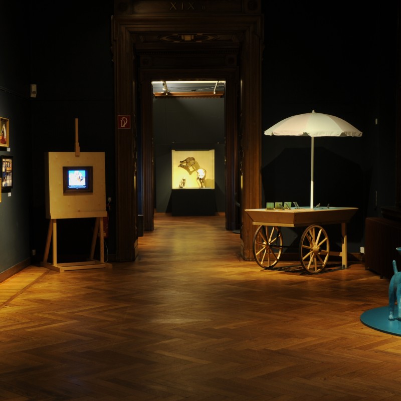 Dimly lit exhibition room with wooden floor, illuminated pictures on the walls, a cart with a white sunshade, and
                                 a blue dog sculpture. (AI-generated alt-text, generated with GPT-4.1-mini)