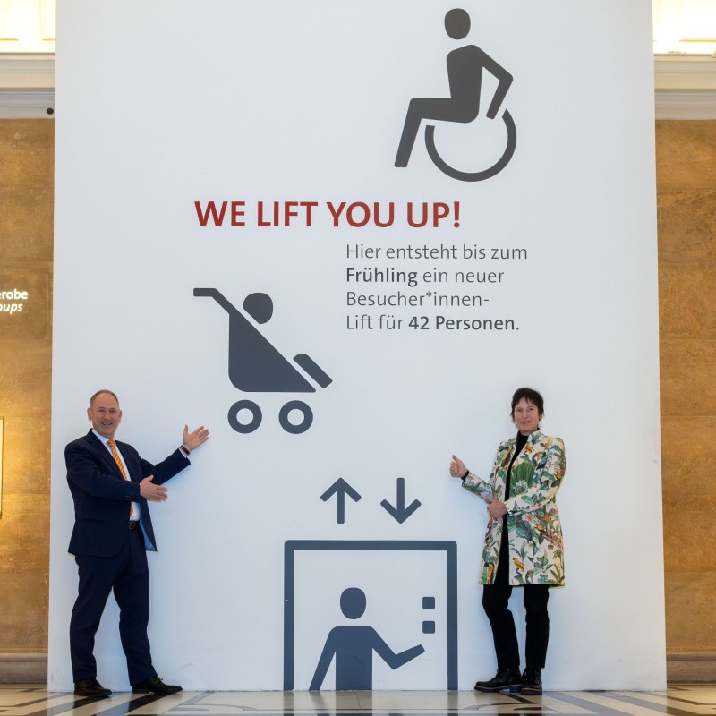 Mag. Markus Roboch und Dr. Katrin Vohland vor einem Plakat im NHM Wien, das den
                                 Bau eines neuen Besucher*innenlifts ankündigt