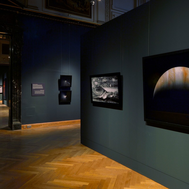 Dimly lit exhibition room with several wall panels showing space motifs, including a large-format planet image with
                                 a shadowed crescent. (AI-generated alt-text, generated with GPT-4.1-mini)