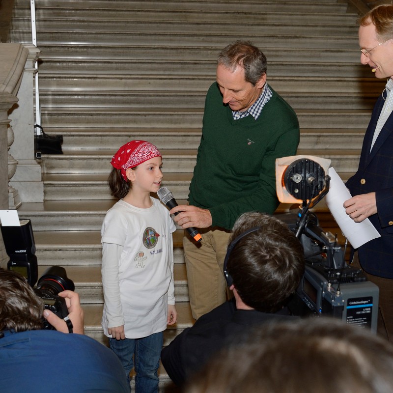 A girl wearing a red headscarf and white long-sleeved shirt speaks into a microphone held by a man in front of a
                                 staircase. Another man with notes stands beside them. Several photographers and camera operators are in the foreground. (AI-generated
                                 alt-text, generated with GPT-4.1-mini)
