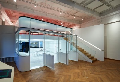 : Modern exhibition space with glass walls, wooden stairs, and view into a bright work area. (AI-generated alt-text, generated with GPT-4.1-mini)