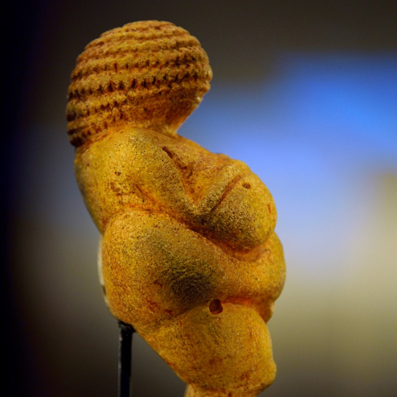 Side close-up of the Venus of Willendorf, a small rounded female figure with prominent breasts and braided-looking
                                 head. (AI-generated alt-text, generated with GPT-4.1-mini)