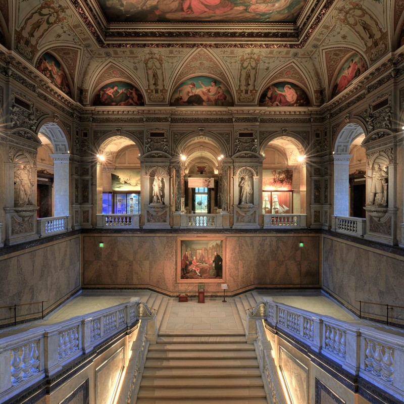 Prächtiges Stiegenhaus mit breiter Marmortreppe, stuckverzierten Bögen, Wandmalereien und reich bemalter Decke.
                                 (KI-generierter Alt-Text, erstellt mit GPT-4.1-mini)