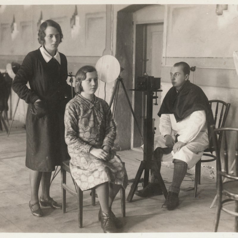 Two women and a man in a room with photographic equipment; one woman sits facing the camera, the others stand and
                                 sit beside her. (AI-generated alt-text, generated with GPT-4.1-mini)