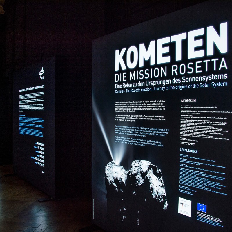 Mehrere
                                 hintereinander aufgestellte, beleuchtete Schautafeln mit Texten und Abbildungen zur Kometenmission Rosetta in abgedunkeltem
                                 Ausstellungsraum. Auf der vorderen Tafel weiße Aufschrift: 'KOMETEN DIE MISSION ROSETTA Eine Reise zu den Ursprüngen des Sonnensystems'.
                                 Unten leuchtendes Foto von zwei Himmelskörpern. (KI-generierter Alt-Text, erstellt mit GPT-4.1-mini)