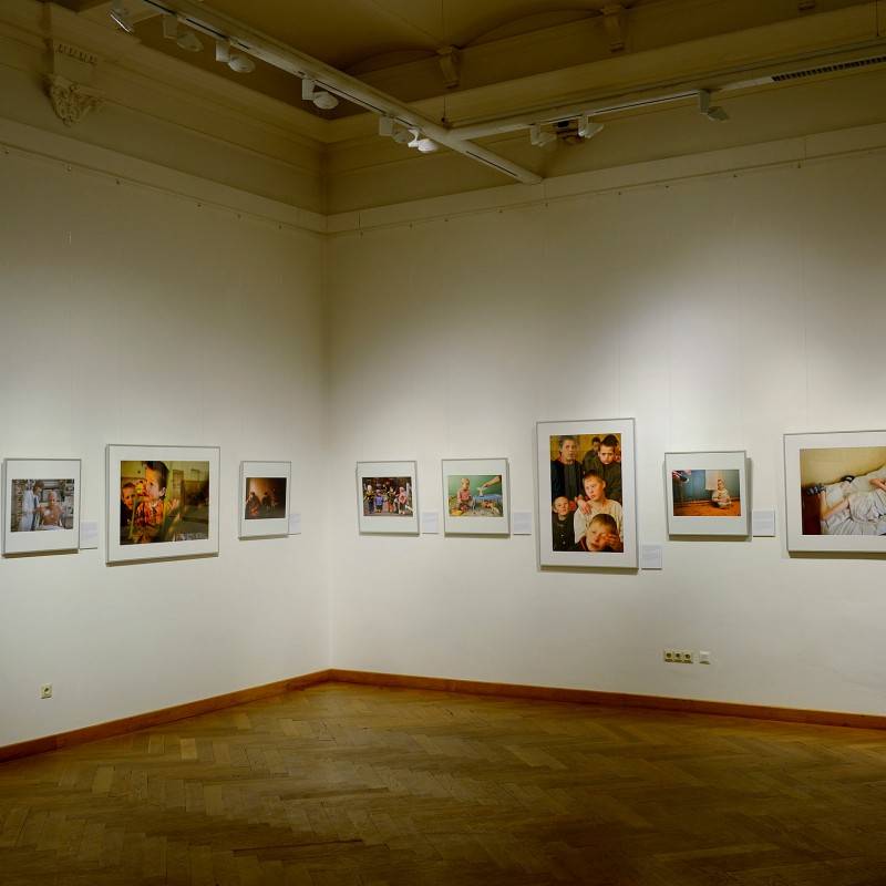 View into an exhibition room with several framed color photographs on white walls, parquet floor, and a light ceiling.
                                 (AI-generated alt-text, generated with GPT-4.1-mini)