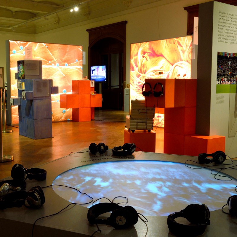 Mehrere Kopfhörer liegen auf einem runden, beleuchteten Tisch. Im Hintergrund sind orange leuchtende Ausstellungselemente
                                 und Monitore zu sehen. (KI-generierter Alt-Text, erstellt mit GPT-4.1-mini)