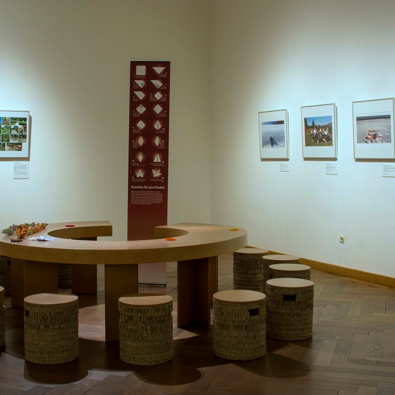 Round wooden table with several stools in an exhibition room, large framed color photographs on the walls. (AI-generated
                                 alt-text, generated with GPT-4.1-mini)