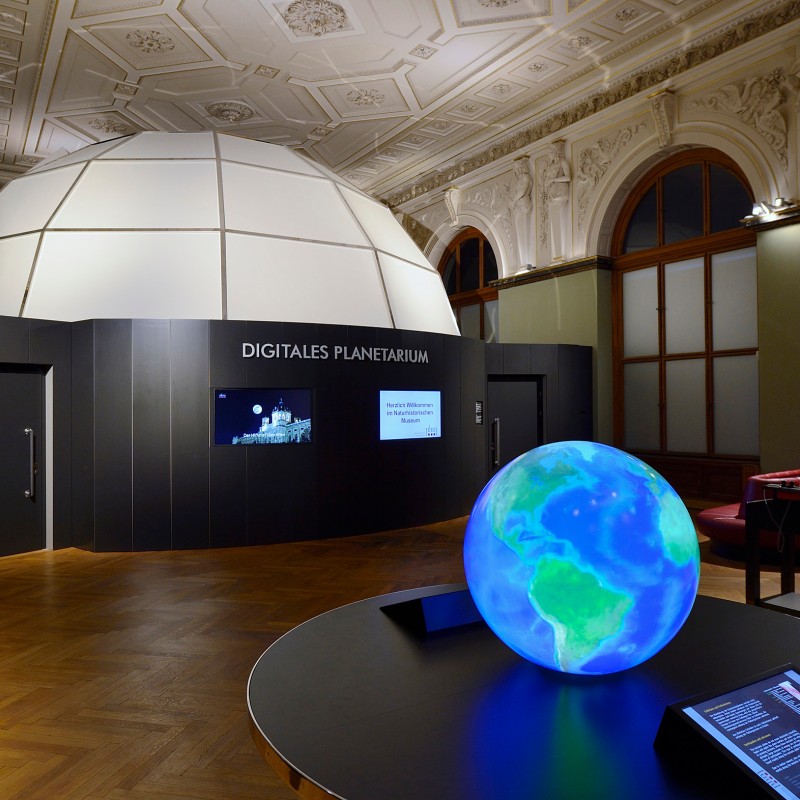 Exhibition room with digital planetarium in the background, illuminated globe and information panels in the foreground,
                                 decorative ceiling. (AI-generated alt-text, generated with GPT-4.1-mini)