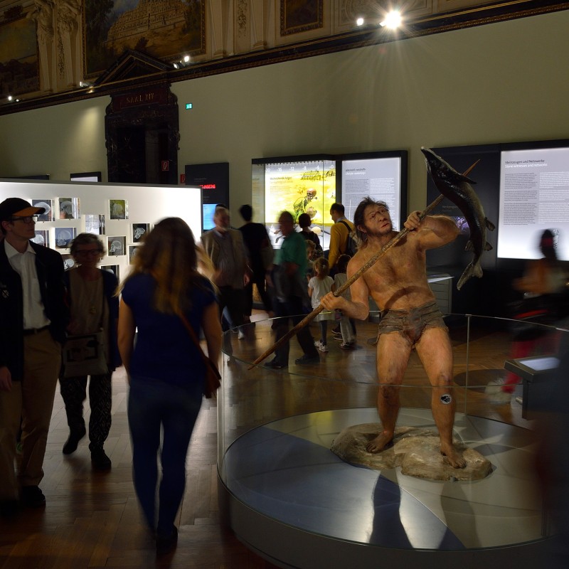 Group of museum visitors passing a life-sized figure of a prehistoric human holding a spear and hunting an animal.
                                 (AI-generated alt-text, generated with GPT-4.1-mini)