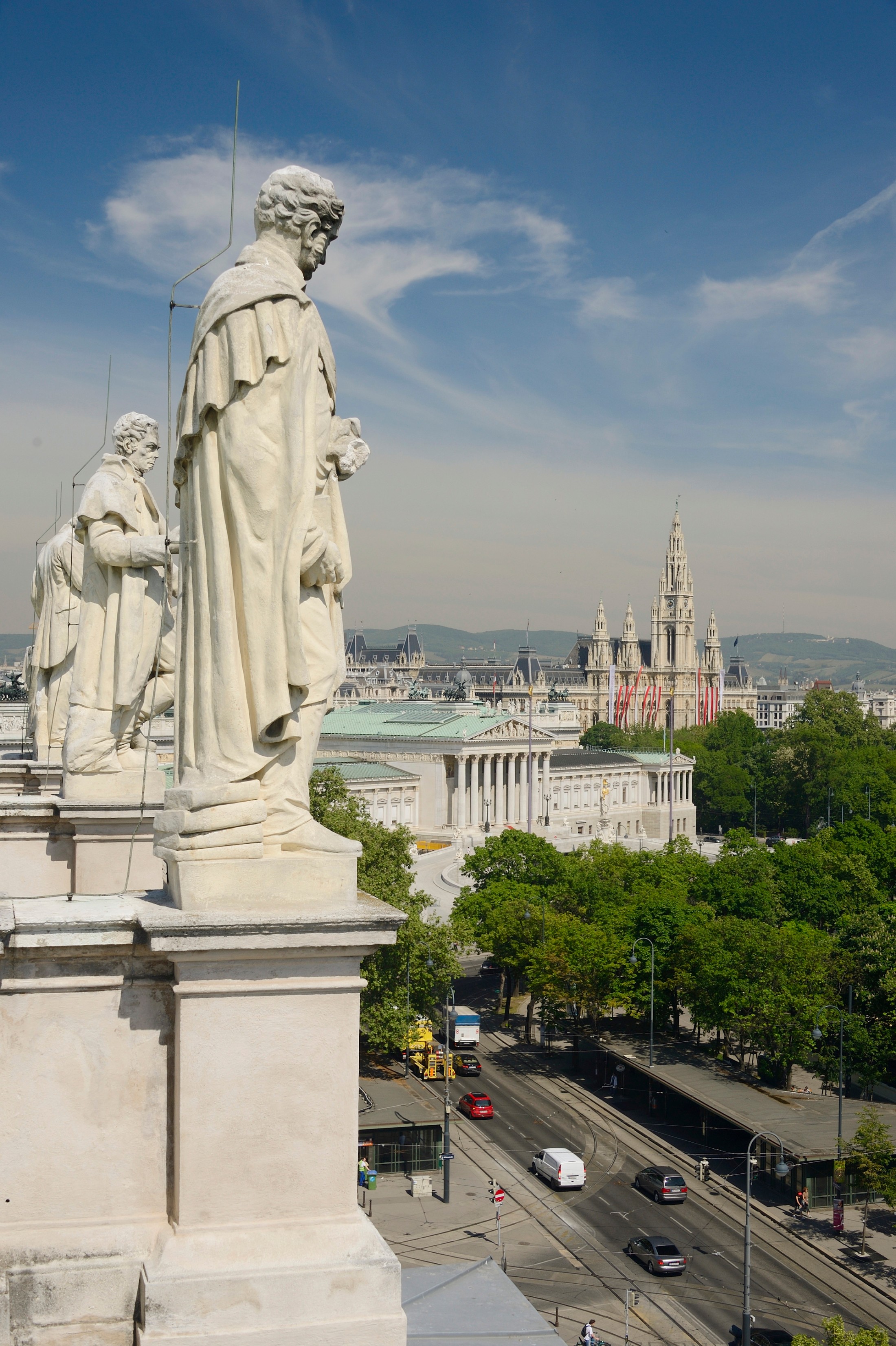 : Steinerne Statuen auf dem Dach des Naturhistorischen Museums Wien, Ausblick auf grüne Baumkronen und das Wiener Rathaus im Hintergrund. (KI-generierter Alt-Text, erstellt mit GPT-4.1-mini)